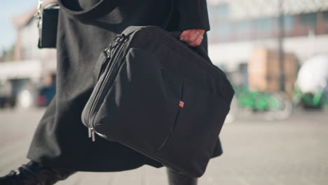 primer plano de una bolsa negra llevada por una persona con un elegante abrigo negro y botas de cuero, caminando con confianza por una calle urbana soleada con arquitectura moderna borrosa y bicicletas verdes en el fondo
