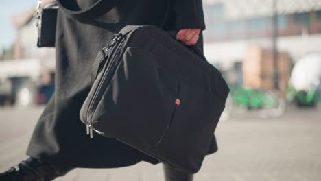 close-up of black bag carried by person wearing stylish black coat and leather boots, walking confidently on sunny urban street with blurred modern architecture and green bikes in the background