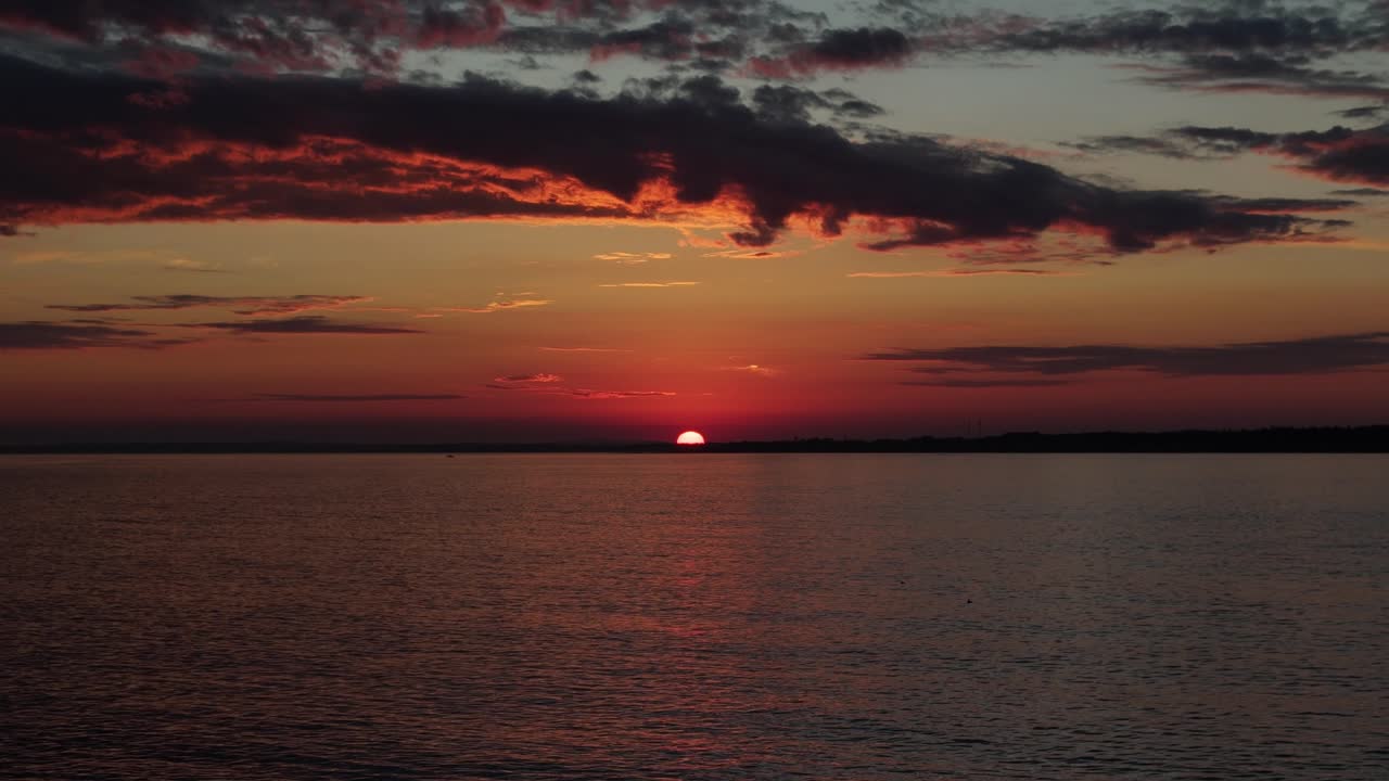 In Maldonado, Uruguay, the last piece of sun disappears over the horizon. Beautiful colors fill the sky, with glowing clouds and soft reflections over the calm water at sunset
