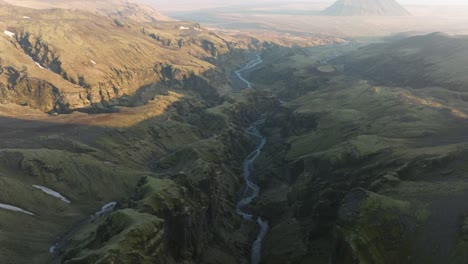 drone view of a vast icelandic canyon with steep cliffs and a winding river, surrounded by rugged terrain and distant mountains