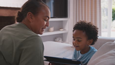 Madre-Con-Hijo-Jugando-O-Transmitiendo-A-Una-Tableta-Digital-Sentados-Juntos-En-Un-Sofá-En-Casa