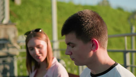 close-up of young man looking aside at friend slightly blurred in the background who is showing him something, serene park setting with trees, iron railing, and lush hill