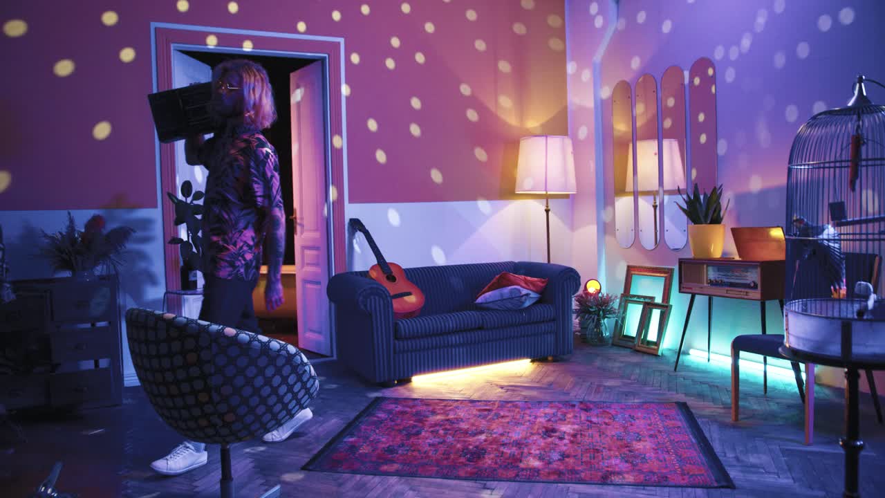 붐 박스를 들고 집에서 복고풍 파티에서 음악의 리듬에 맞춰 걷는 긴 머리를 가진 젊은 세련된 백인 남자