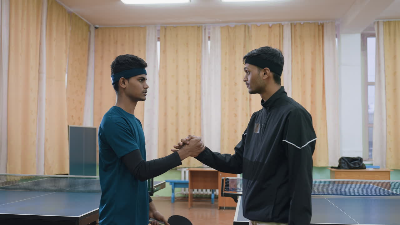 Two tennis players shake hands near table tennis table indoors, demonstrating sportsmanship, respect, and camaraderie after practice session, wearing athletic outfits