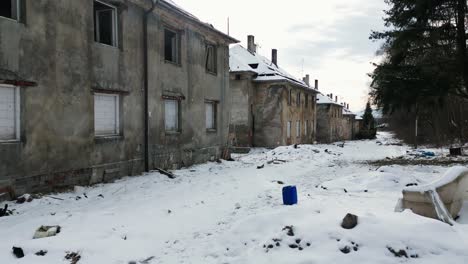 series of neglected, snow-covered buildings with boarded-up windows, surrounded by trees and scattered debris