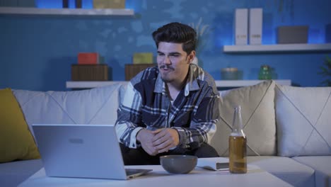 un joven viendo una película por la noche en su portátil en casa. está bebiendo alcohol.