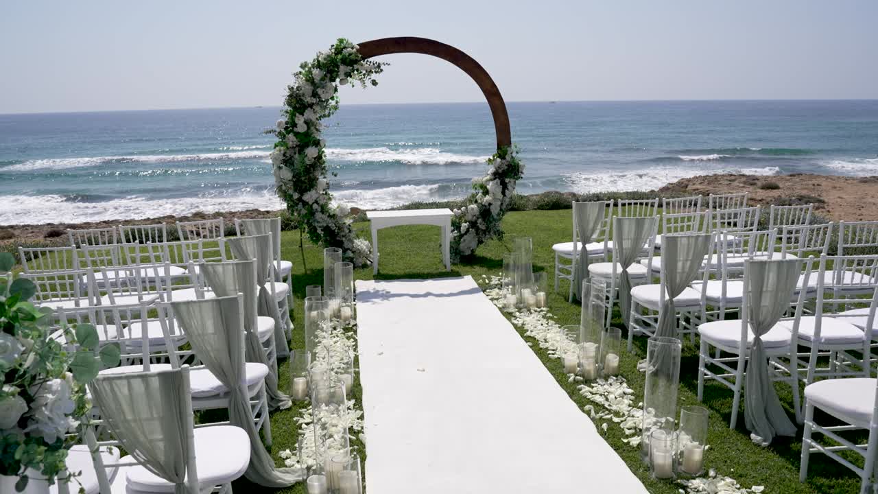Un allestimento per un matrimonio all'aperto è organizzato in riva al mare. Sedie bianche sono allineate di fronte a un arco fiorito. Il sole splende luminoso sull'oceano e sul paesaggio costiero.