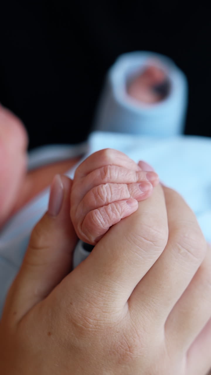 Tiny newborn child wearing blue romper in the parent's hands. Loving mother is holding cute fingers and kissing baby on the head. Close up. Vertical video