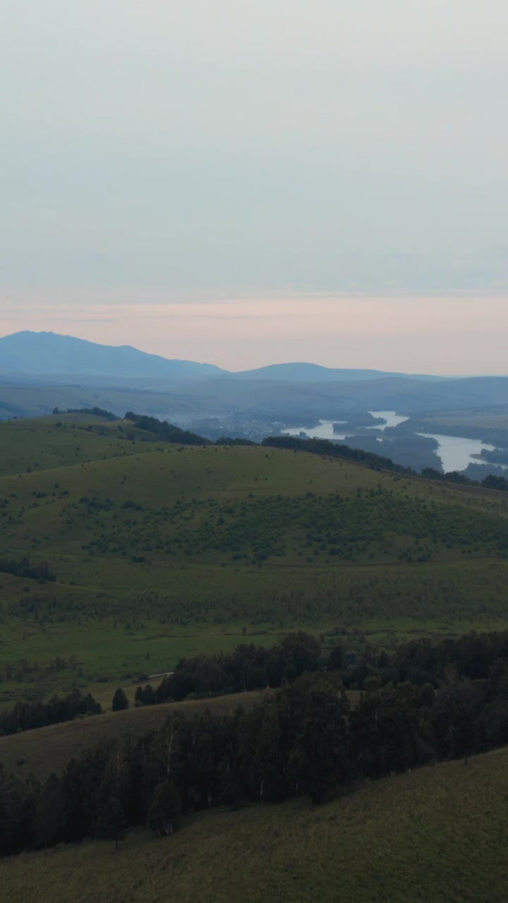 Ampia veduta cinematografica di dolci colline verdi e un fiume tortuoso al tramonto sui Monti Altai, Russia, con una leggera foschia e creste montuose distanti sotto un cielo calmo.