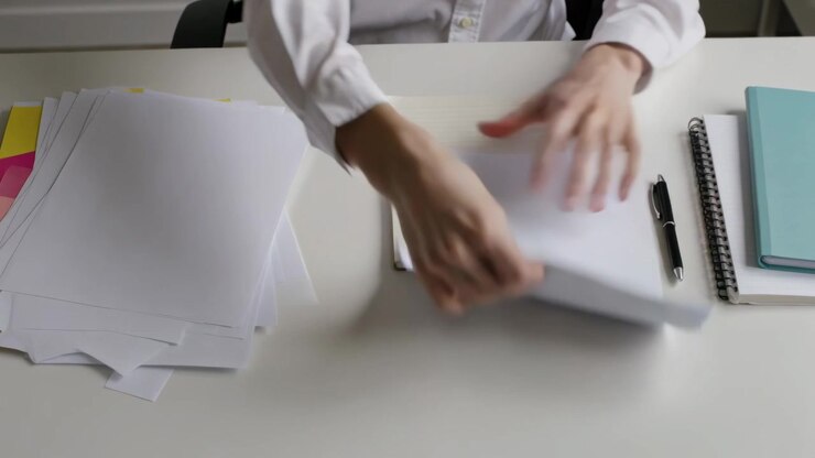 Spreading stacks, person in white shirt sorting loose sheets at desk, preparing notebook and pen