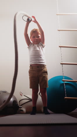 blond little boy waves battle rope standing near gym equipment. strong toddler tries effective exercise for strengthening body at home slow motion