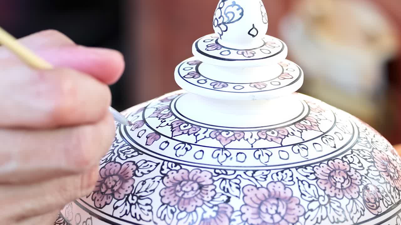 Close-up of an artist painting floral designs on a ceramic lid using a fine brush.