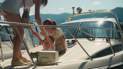 mother pulls out little boy of boat hatch with girl in sunny city port on summer day. cute children with woman leave moored yacht against mountains