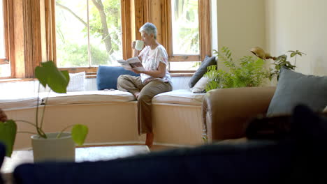 happy senior biracial woman drinking tea and reading book at window at home, slow motion