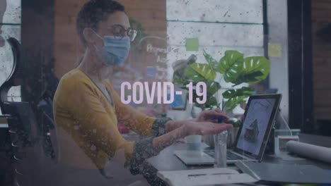 Covid-19-text-and-grunge-effect-against-woman-wearing-face-mask-sanitizing-her-hands-at-office