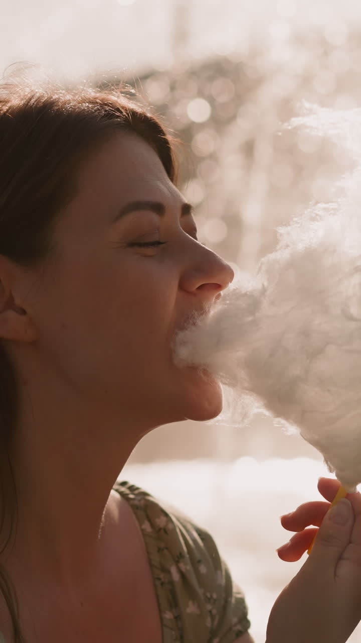 Attractive woman eats cotton candy against blurry fountain at sunset closeup slow motion. Enjoying dessert in amusement park. Lady carefree childhood memories