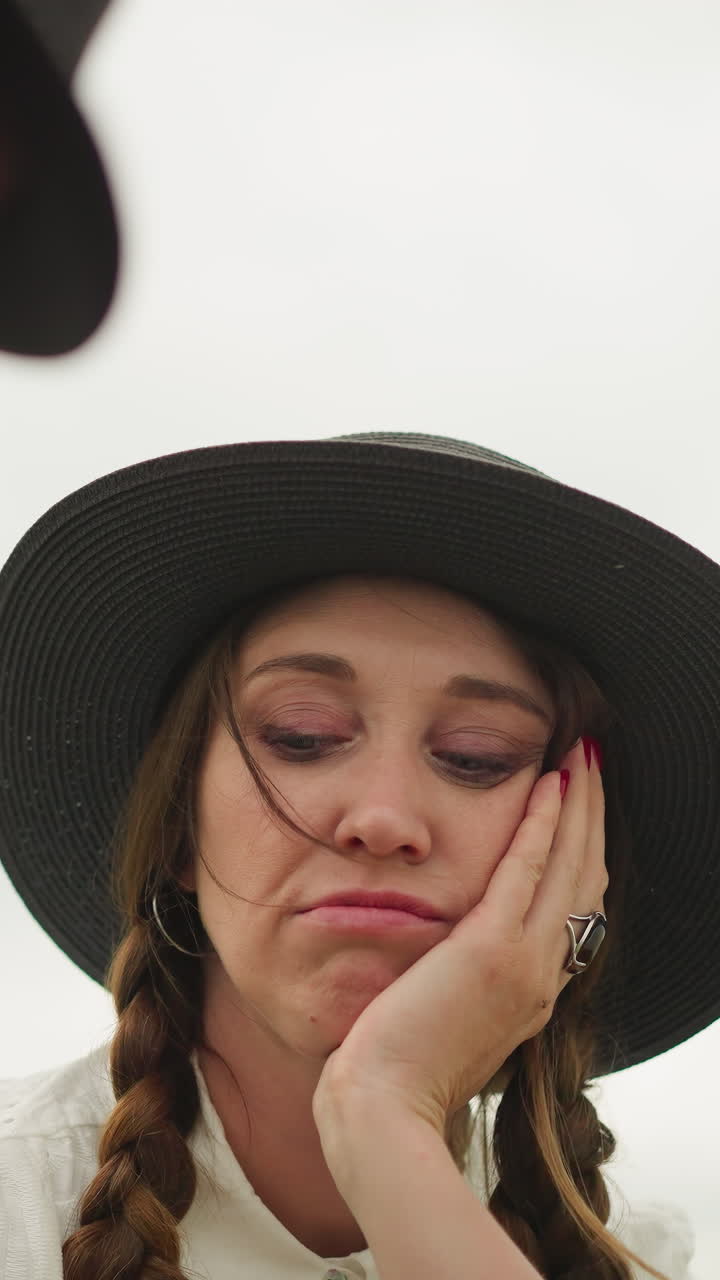 un primer plano de una mujer con un sombrero y un vestido blanco, mostrando una expresión agotada con la mano en la cara. en el fondo borroso, un hombre es parcialmente visible