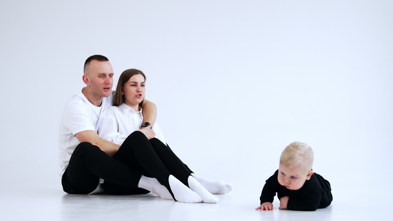Young parents sit on the floor looking at their adorable crawling child. Mom and dad talk and laugh to their baby. White backdrop.