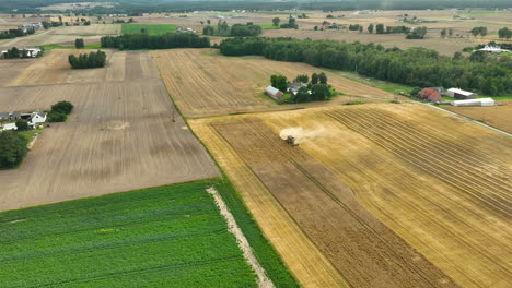 aerial view of a combine harvester working on a wheat field near a farm, with visible dust trail and green fields surrounding the area