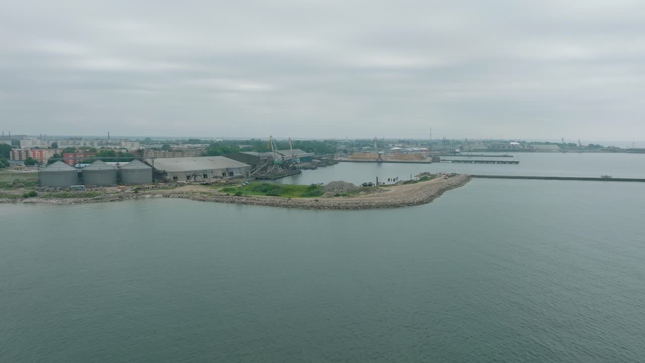vista aérea de las grúas portuarias y los muelles de carga vacíos en el puerto de liepaja, la ciudad de liepaja en el fondo, día de verano nublado, disparo de avión no tripulado en órbita a gran distancia moviéndose a la izquierda