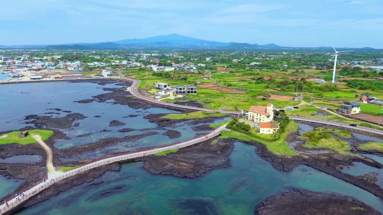 Paralaks udara di atas Taman Singyemul Pulau Jeju menampilkan jalan setapak melengkung di atas pantai vulkanik hitam, bangunan bergaya Eropa, jalan Sinchang, dan gunung Hallasan