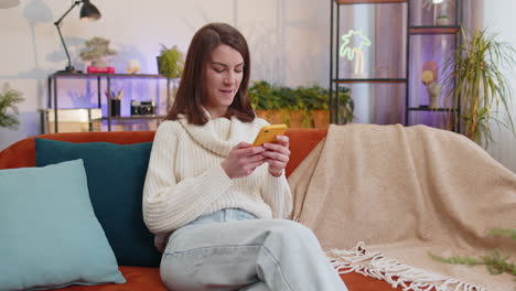 Young-woman-sitting-on-sofa-at-home-using-smartphone-share-messages-on-social-media-application