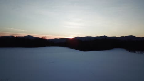 Drone flight over a snowy field in late winter, with the setting sun in the background over the mountains, with trees and forests coming into view.