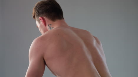 side profile of determined shirtless high school boy with geometric neck tattoo working out by pulling heavy resistance rope in sunlit room, sweat bead visible, muscle tone defined