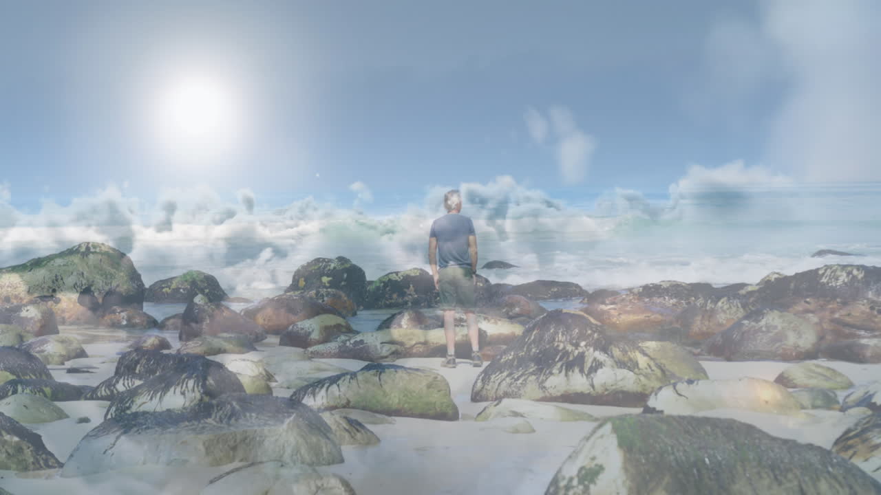 кавказский пожилой мужчина стоит на пляже и смотрит на море, над быстро движущимися облаками