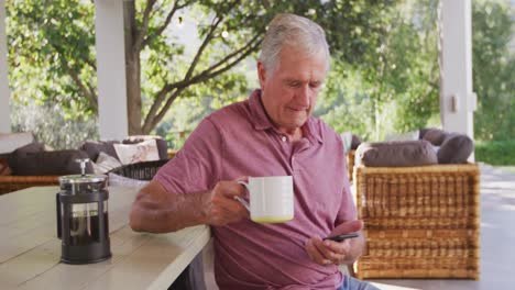 Senior-Caucasian-man-using-his-phone,-drinking-a-beverage-in-the-garden