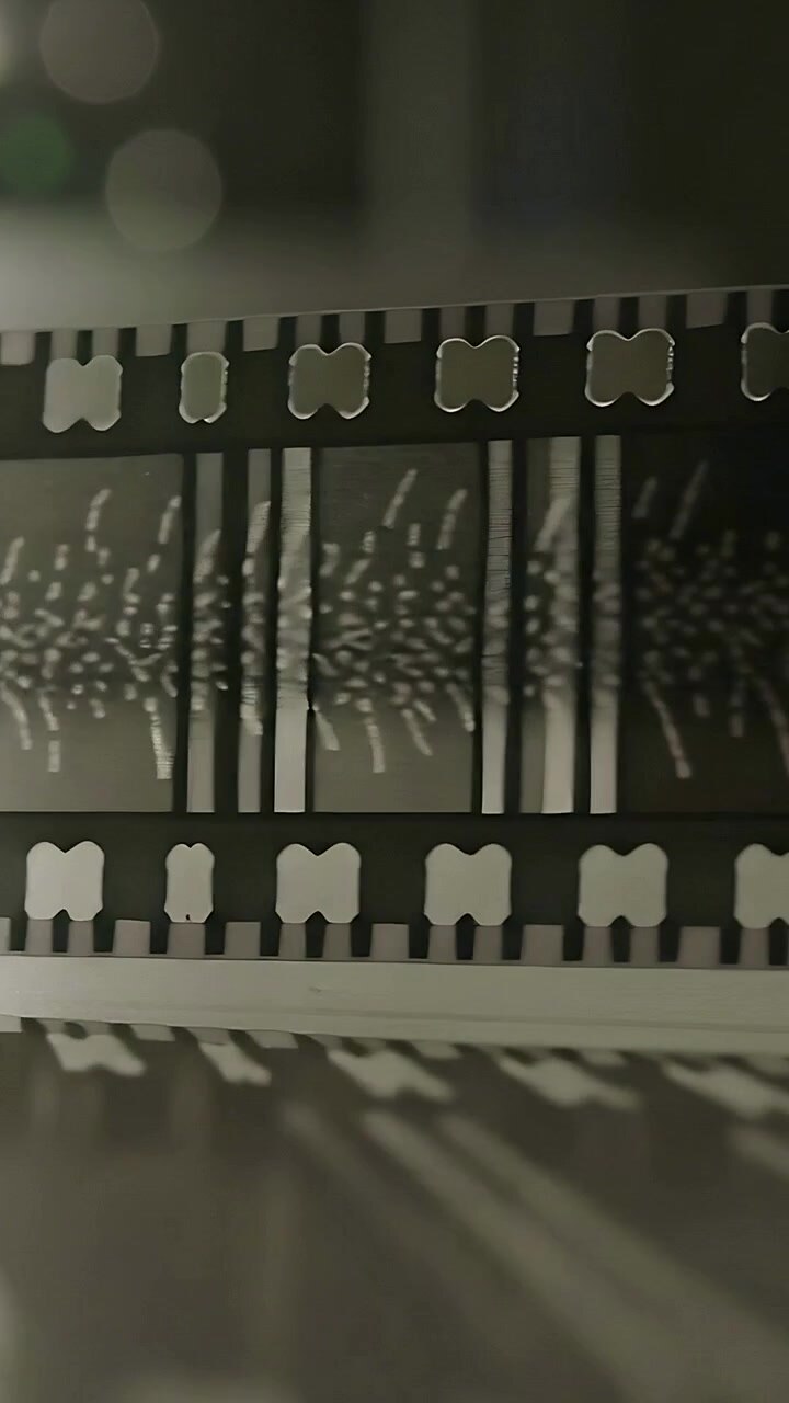 Video verticale: Motore che tira una striscia di pellicola perforata sopra un tavolo luminoso, rivelando granelli luminosi
