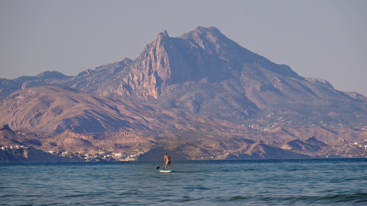 Lone paddleboarder on calm water beneath a towering, craggy mountain in Alicante, Spain