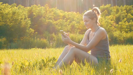 happy young woman sitting on green lawn and uses smartphone on scenic field at sunset background