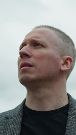 a close view of an aged white man wearing a grey suit and a black polo shirt, turning his attention to the left with a focused expression, with an overcast sky
