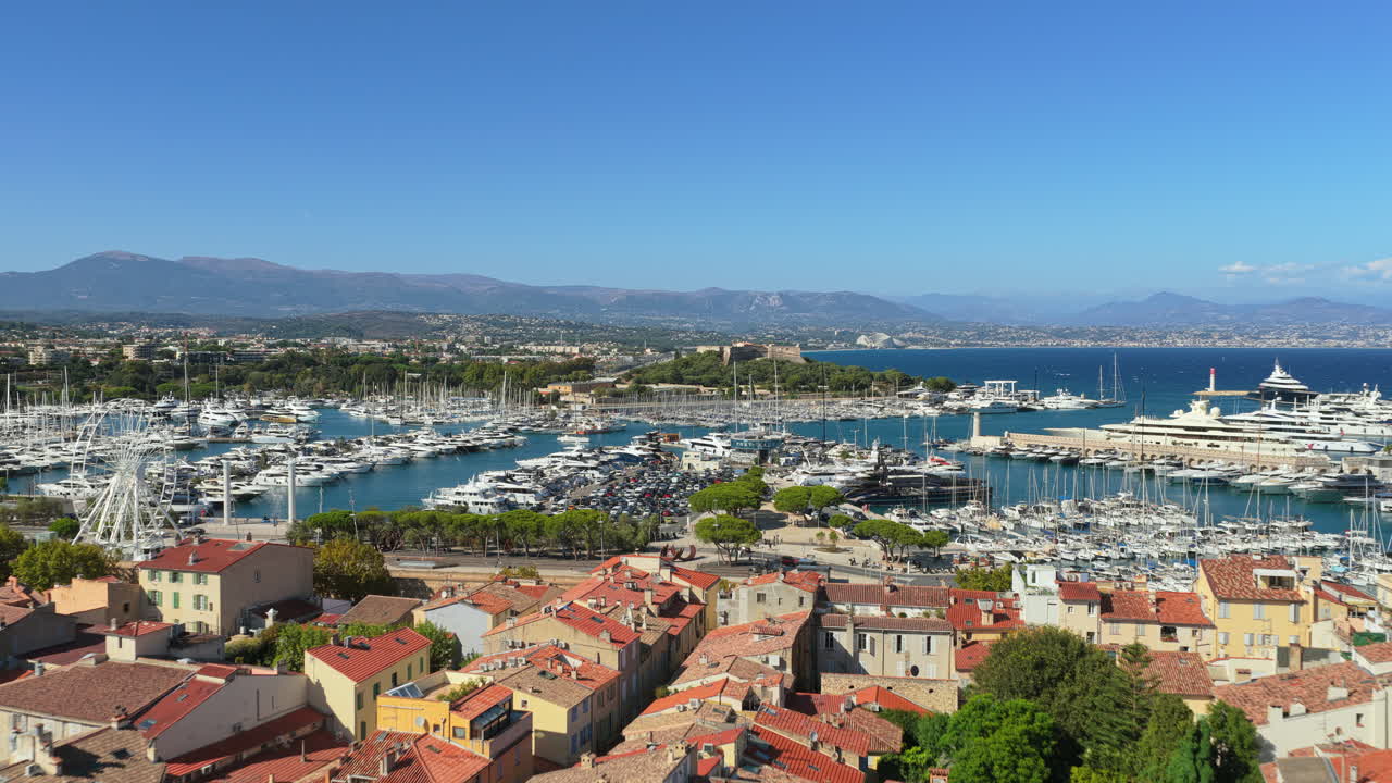 Aerial drone view of the marina filled with luxury yachts and sailboats, with the Ferris wheel, coastline, and Fort Carre in the background, Antibes, France