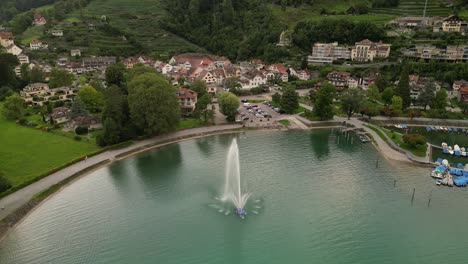 bird view flying towards a fountain splashing water in air placed inside a lake cars parked along the shore and brown houses standing under the hills beautiful architectures peaceful environment