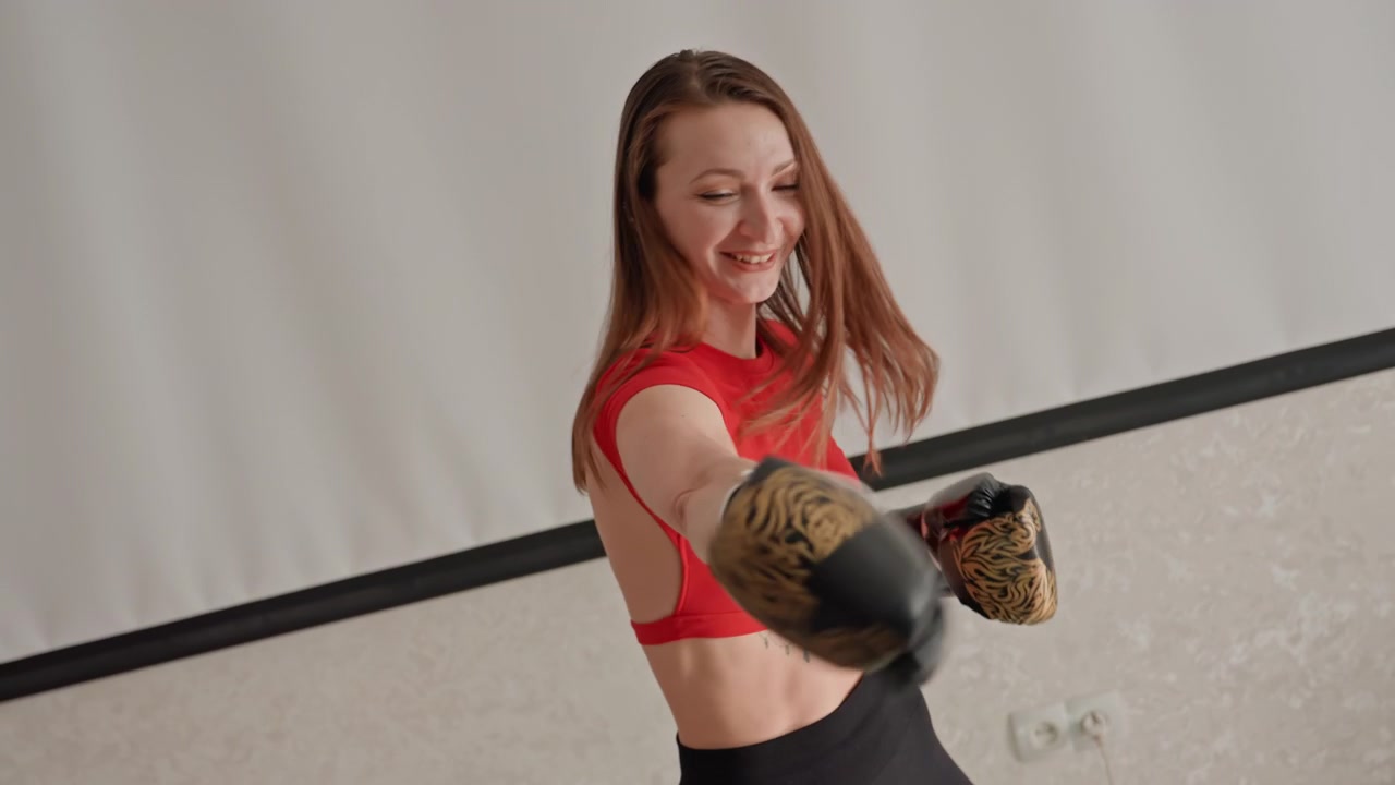 Pratica di boxe a casa in piedi, donna bianca che esegue esercizi tecnici, sorridente con crop top rosso e leggings neri, guantoni con motivo dorato, ritmo rilassato, sessione di fitness casalinga accessibile, equilibrio.