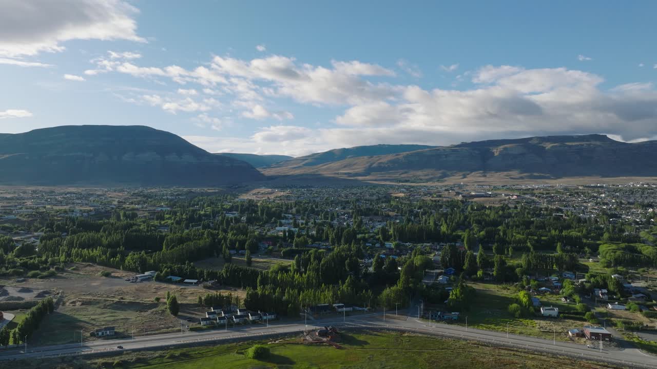 Aerial view of El Calafate town nestled in Patagonian valley with dramatic mountain backdrop. Residential neighborhoods spread across green landscape under expansive cloudy sky.