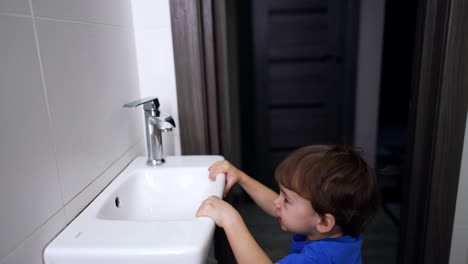 dark-haired toddler in blue t-shirt enters the bathroom. kid approaches the sink and opens the tap.