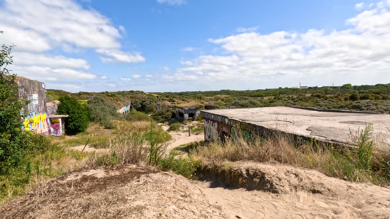 Camera slowly pans right over a sunlit, graffiti-covered historic WWII bunker surrounded by sand, grass, and shrubs under a partly cloudy sky
