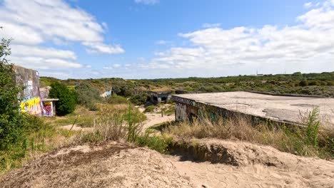 camera slowly pans right over a sunlit, graffiti-covered historic wwii bunker surrounded by sand, grass, and shrubs under a partly cloudy sky