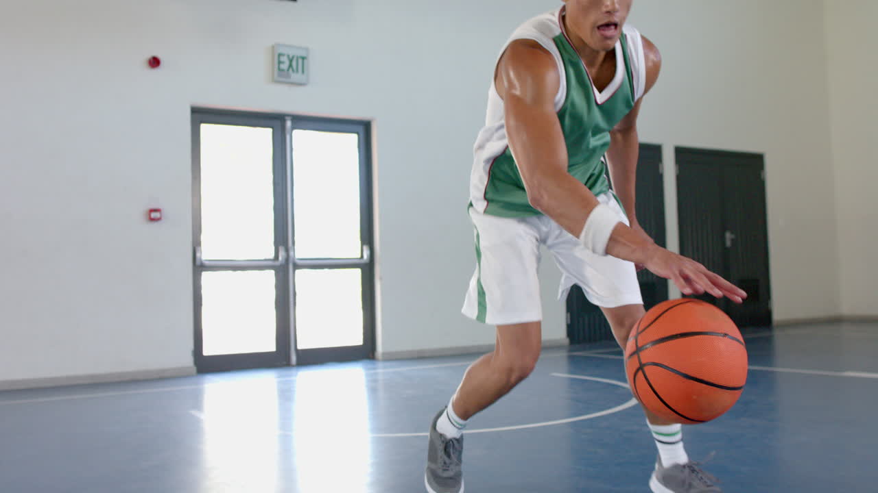 un joven biracial con una camiseta verde dribla una pelota de baloncesto en un gimnasio