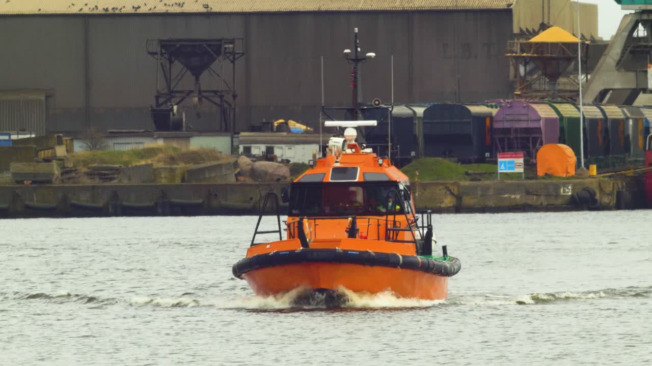 barco piloto del puerto naranja saliendo del puerto de liepaja, vagones de carbón estacionados en el fondo, día nublado de primavera, plano medio desde la distancia