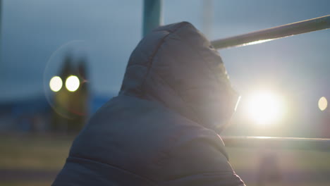 close-up of a person in a hooded jacket resting on an iron bar with a soft light reflection in the background shinning from the distance