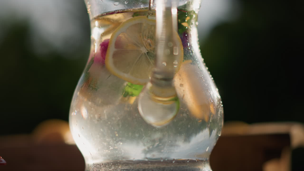 Primo piano di una brocca scintillante con una fetta di limone e un cucchiaio che mescola, bolle che salgono attorno agli agrumi, bagliore del sole attraverso il vetro, condensa sulla brocca, movimento dinamico di versamento, rinfresco effervescente per l'estate.