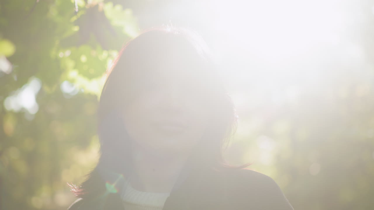 Portrait of fashionable girl standing among leafy trees, eyes lowered, sniffing dry leaf with soft smile as warm sun beam paints face, serene forest bokeh around, calm mood during quiet autumn walk