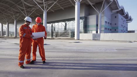 Two-construction-workers-in-orange-uniform-and-helmets-meeting-each-other-at-the-bulding-object,-shaking-hands-and-examining-the-constructed-building-together.-Teamwork-concept