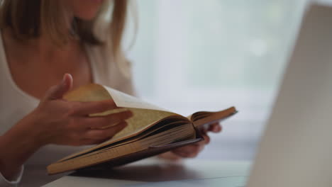 una mujer rubia da la vuelta a las páginas de un libro viejo sentada cerca de una computadora portátil