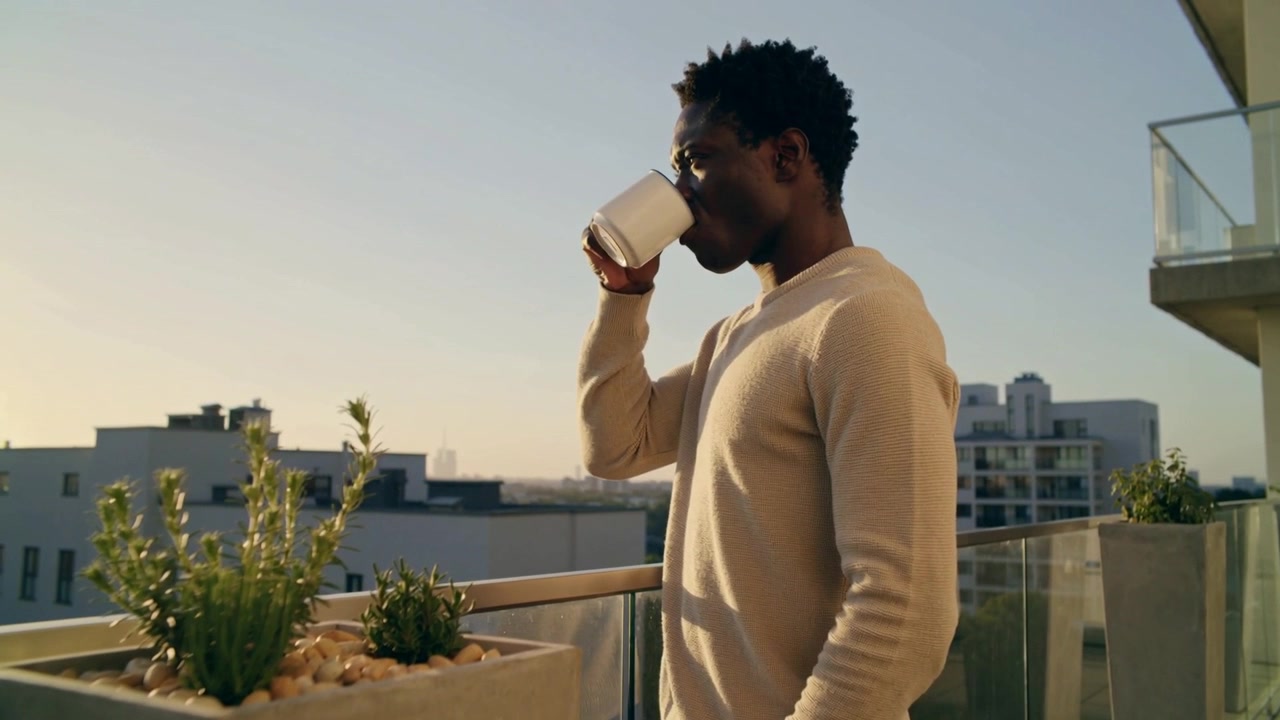 La luce del sole illumina un uomo con un maglione color crema che solleva una tazza bianca e sorseggia sul balcone, vicino a una fioriera di erbe aromatiche.