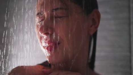 positive woman with water drops on face in shower unit closeup. pretty lady enjoys warm jets washing body in home bathroom. relaxation in washroom
